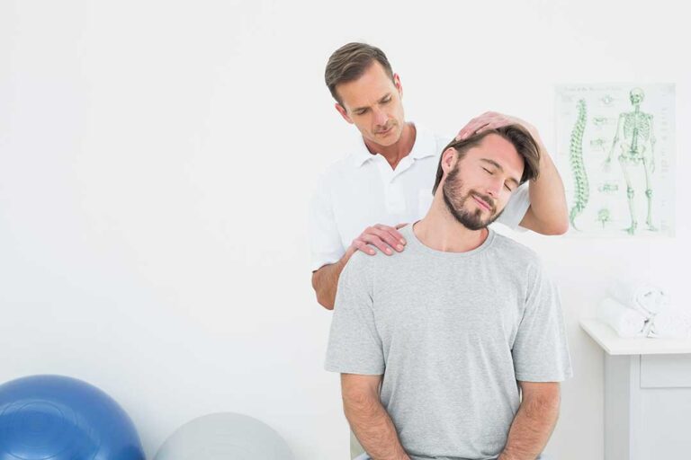 A person sits on a couch with a hand on their lower back, looking puzzled as they rub their shoulder.